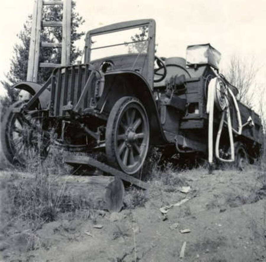 A truck used to fight forest fires. Pictured on the left side of the truck is a hose as well as an axe. The description on the back of the photograph says "1921 misc. white truck, Bovill."