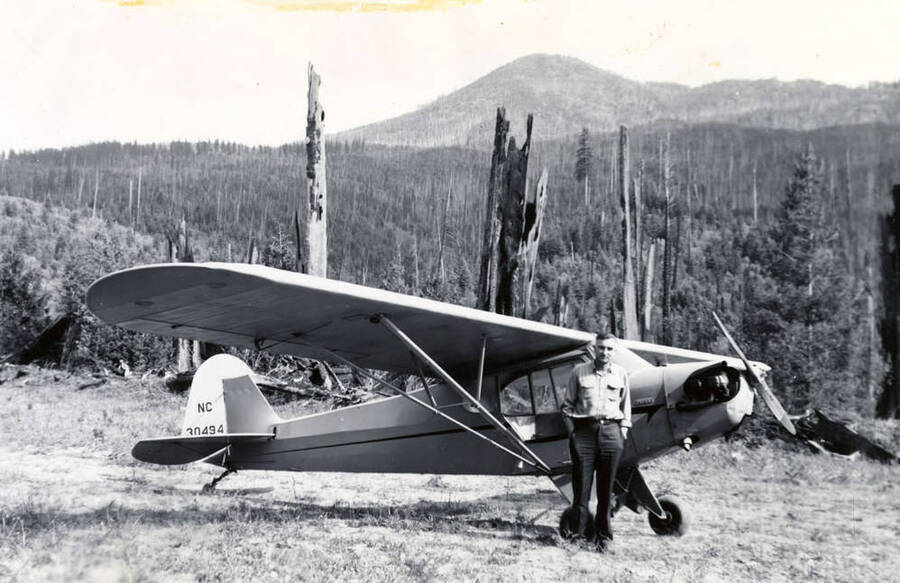 A man stands in front of a fire patrol plane. Behind him and plane, two trees bare the mark of recent forest fires. The tail number on the plane is NC 30494.