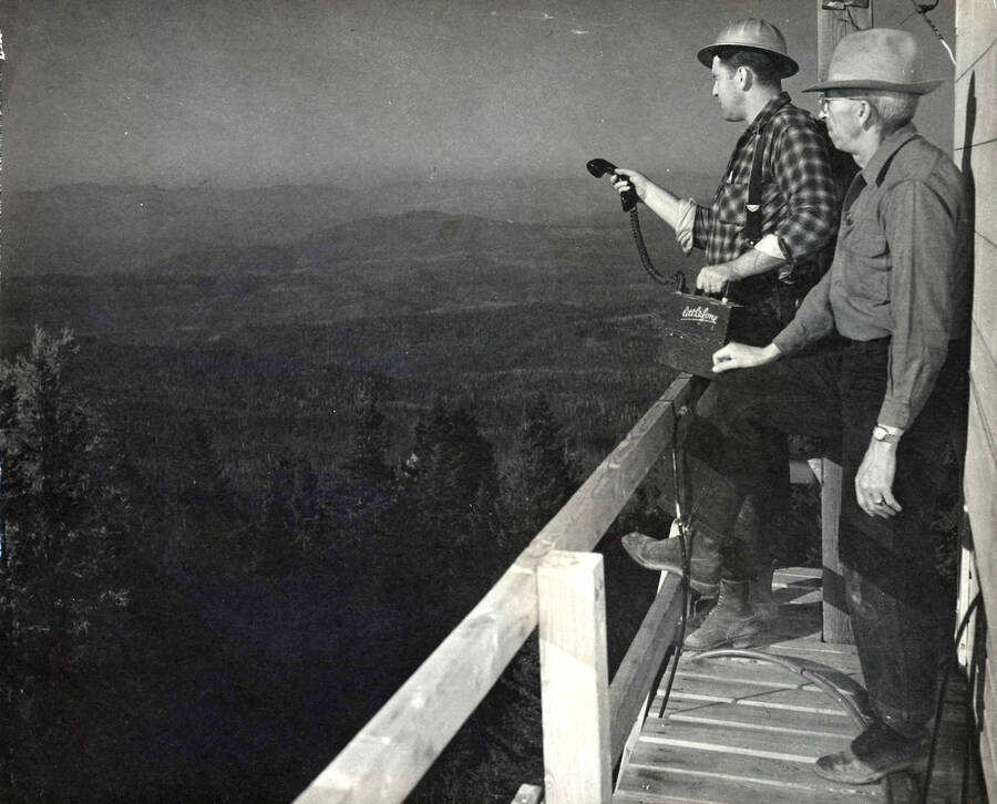 Two men look out over the forest from a fire lookout station. The man in the hard hat is holding a portable phone device, which the box is labeled 'Littlefone'