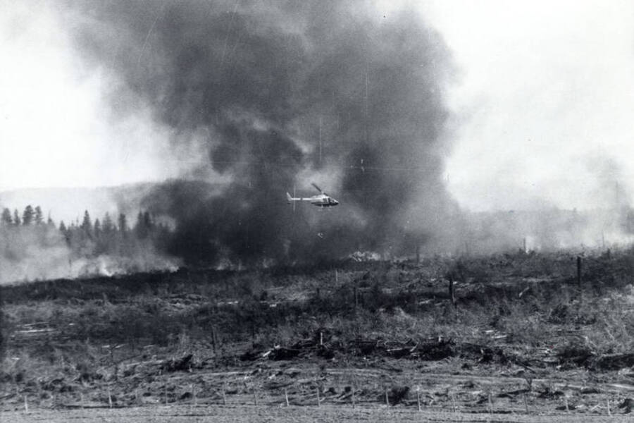 A helicopter flies through smoke during a controlled burn, using a drip torch to start more fires. The land below the helicopter is burned.