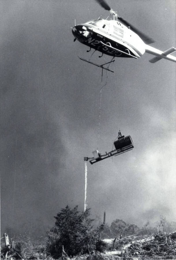 A helicopter uses a drip torch to light cleared brush and logs on fire. Written on the belly of the helicopter is N2510.
