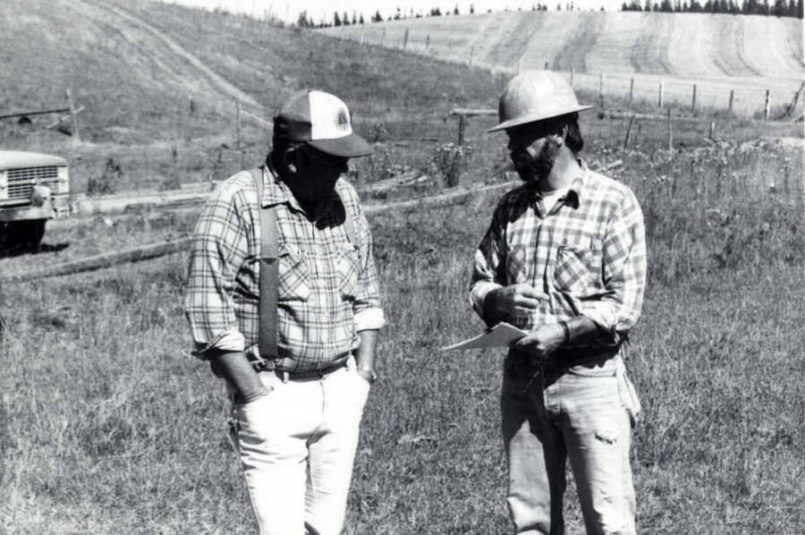 Two men discuss the plan for the controlled burn. In the background, a field has been harvested. On the left hand side of the photograph the front part of a truck is visible.