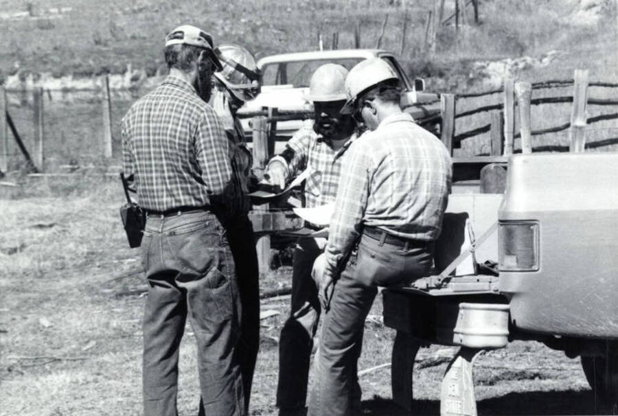 A group of men discuss the plan for the controlled burn. Two of the men are standing while the other two are sitting on the bed of a pickup.