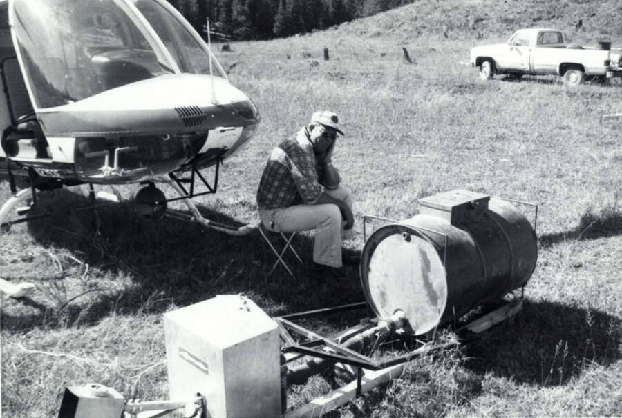 A man sits in front of a helicopter and the drip torch. In the background is a pickup.