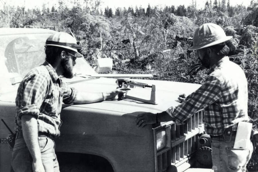 A man weighs a piece of wood on the hood of a pickup truck while another watches. Behind them is brush and limbs gathered in preparation for a controlled burn.