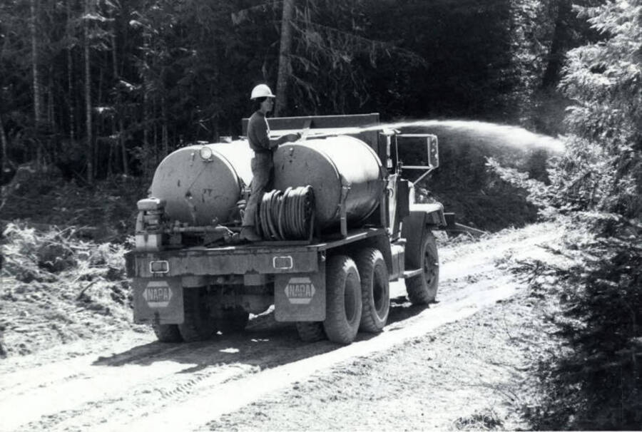 A man rides on the back of a water truck spraying down trees and the ground as part of the management for the burn.
