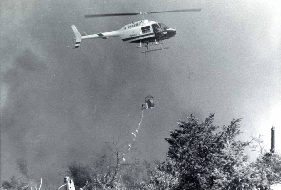 A helicopter uses a drip torch to light brush and limbs on fire for a controlled burn. On the side of the helicopter it says 'Potlatch'