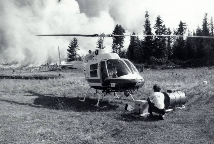 The helicopter used in a control burn. In front of it is the drip torch used to light the fire. Kneeling next to the drip torch is a man. In the background a large cloud of smoke can be seen.