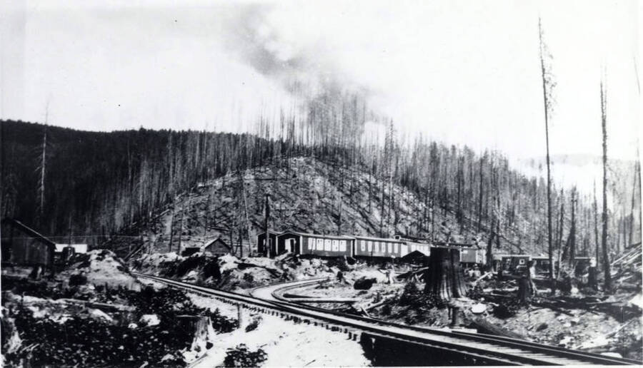 Camp D - Upper Basin near Cameron Creek. In the foreground, train tracks can be seen while in the background, smoke from a forest fire can be seen.