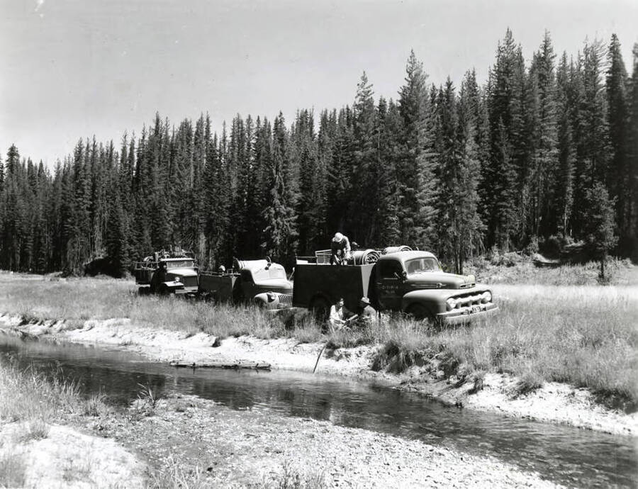 Men work to fill the water trucks used to fight forest fires from a stream. The description on the back says "Filling TPA [Timber Preservation Association] tankers. AB Curtis photo file."