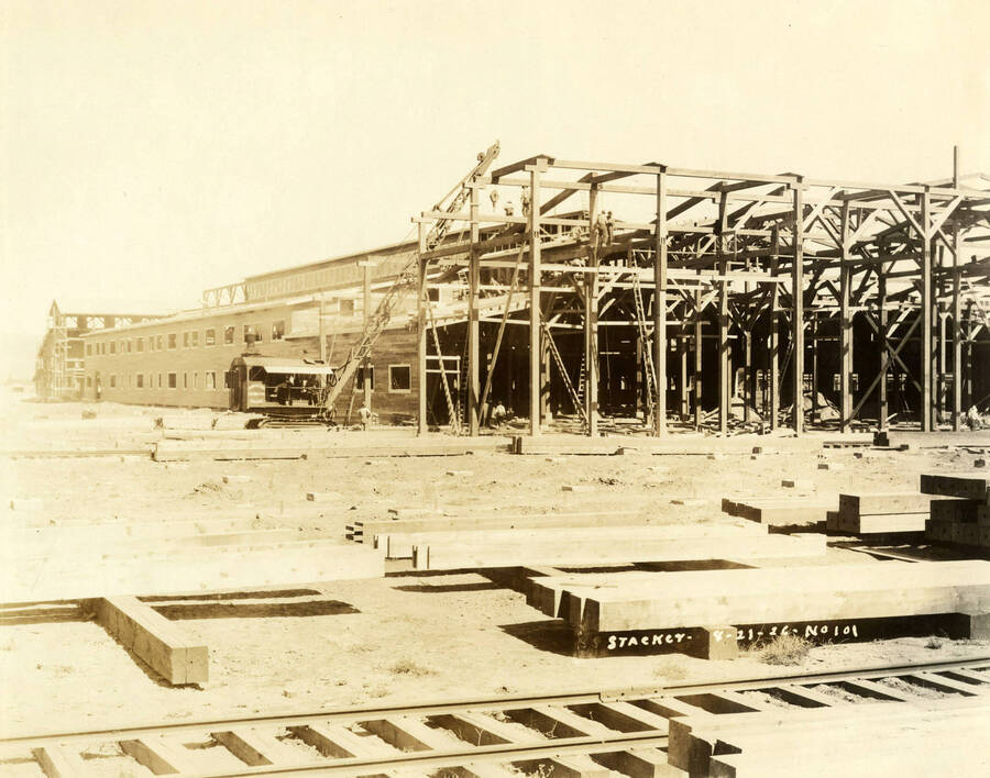 A crane is use to help construct a building at the Lewiston Mill. In the foreground of the photograph lie planks of lumber as well as railroad tracks. Written on the photograph is 'Stacker 8/21/1926 No. 101'