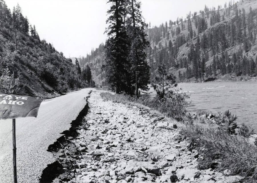 The road that was damaged from floodwaters. On the left side of the photograph  is a sign that reads "state of Idaho." A river can been seen on the right of the photograph.