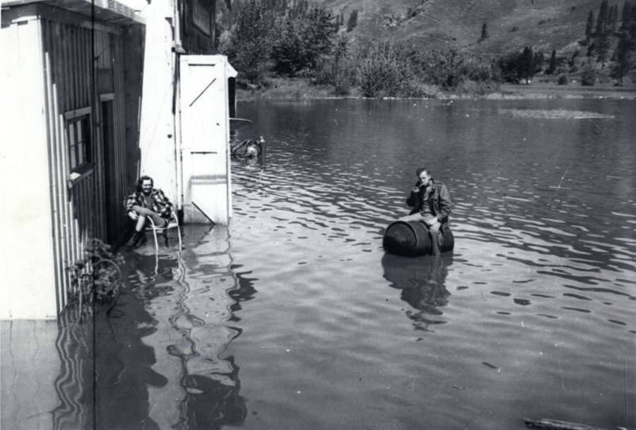 A woman sits with her legs up over the side of a chair while a man sits on a barrel in the middle of the floodwaters.