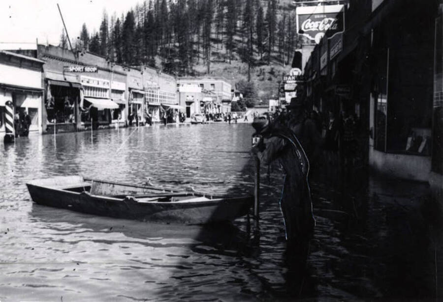 A man stands next to a boat floating in the main street flooded. In the upper part of photograph a sign for Coca-Cola is seen.