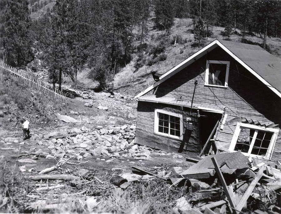 A man walks away from a house damaged by the flood. Rocks are piled to the left of the house while in the foreground of the picture debris is pictured.