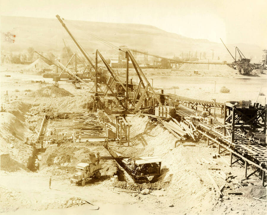 Machines and cranes work to construct the dam. At the bottom of the picture, a crane dumps rock and dirt into the back of a dump truck. Written on the photograph is 'Dam 8/22/1926 No. 104'