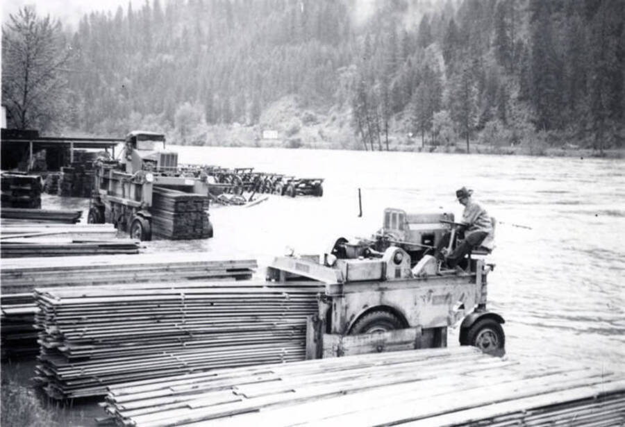 A man drives a piece of lumber machinery to move stacks of lumber while the river floods behind him