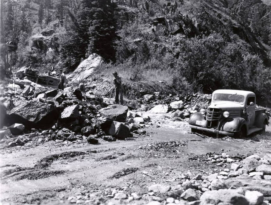 Two men work on clearing a road washed out in the 1948 flood. A pickup truck sits as water runs in front of it.