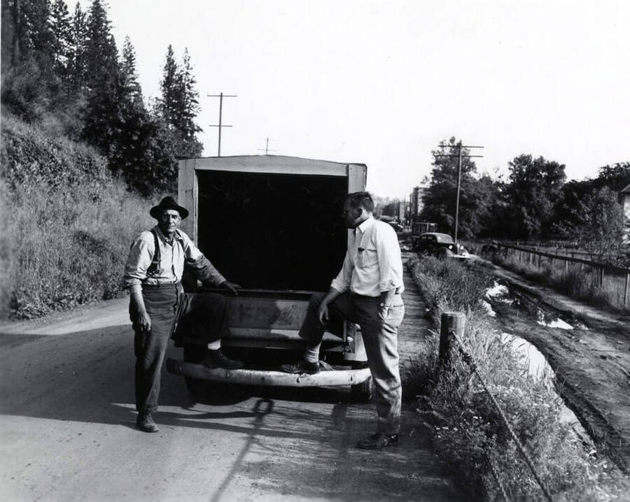 Two men stand  with one foot on their truck on a road. Another car is in the background.