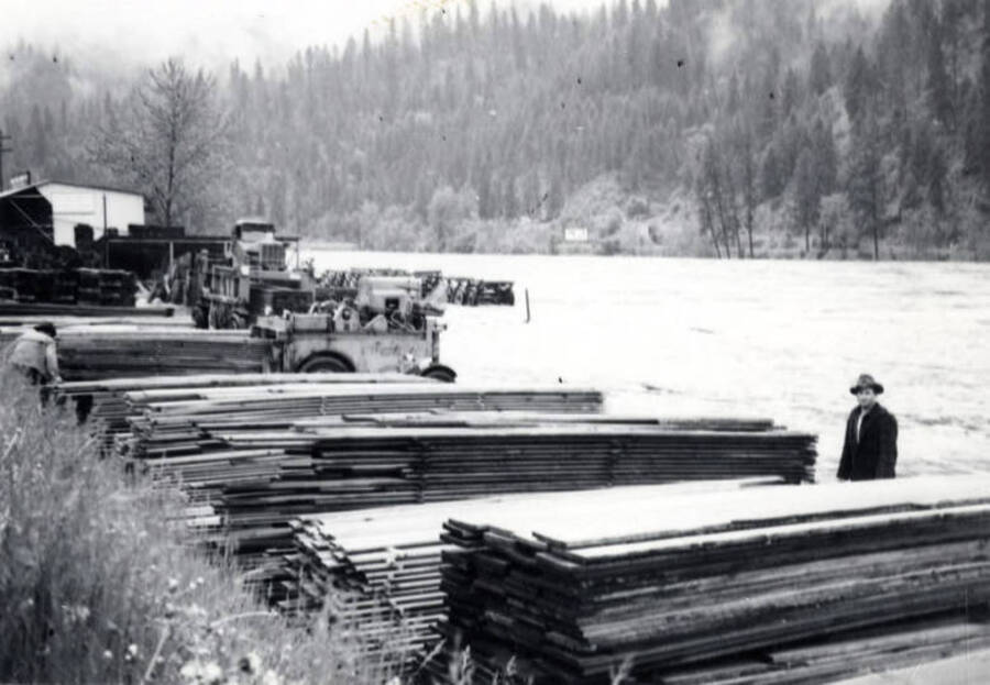 A man stands in front of stacks of lumber while the river behind him floods. In the background are two pieces of mill machinery.