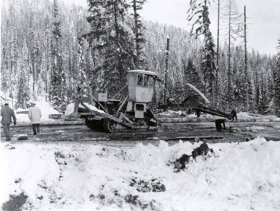 Two men walk behind a piece of lumber machinery. Written on the back of the photograph is 'Cherry Picker" - Robinson Cr. Landing - 1/13/1960."