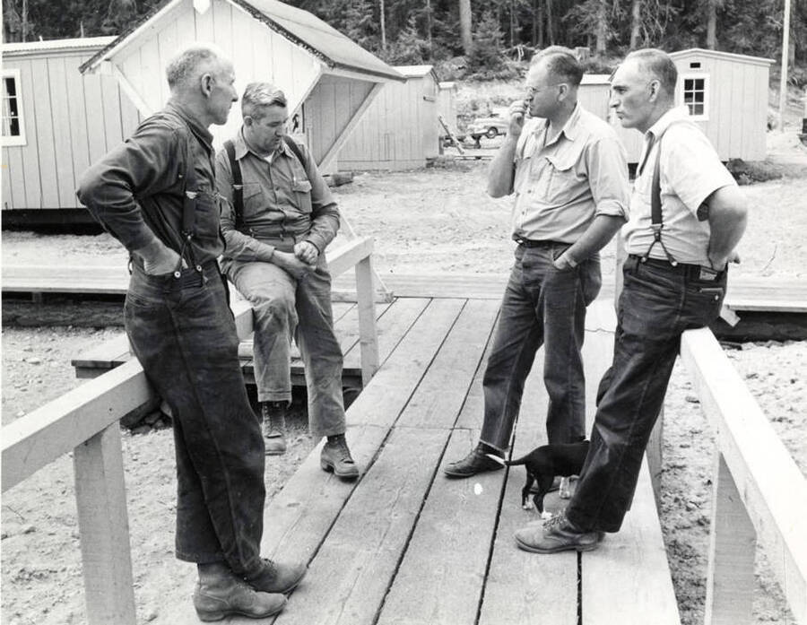 Four camp men stand around talking. Written on the back are the names of the men. They are from left to right: Dooley Cramp, Earl Ritzheimer, Bert Curtis, and Dwayne Space. At Mr. Curtis's feet is a small black and white dog.