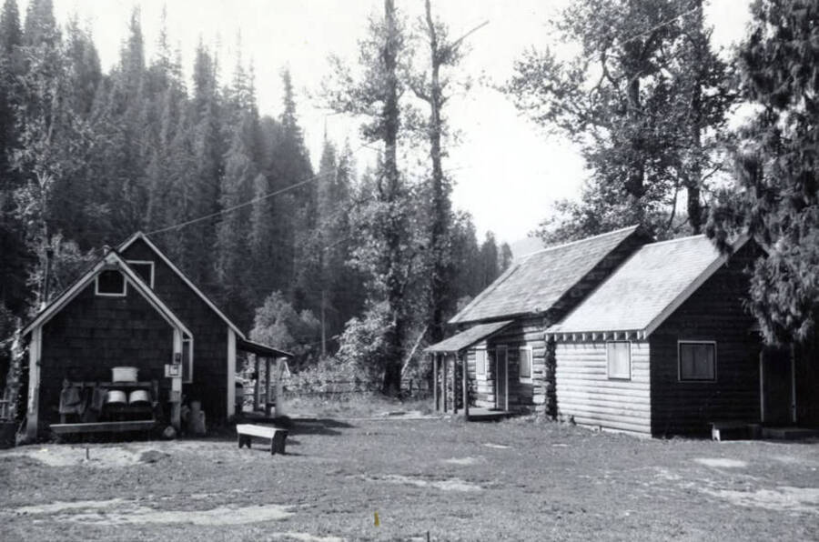 A log cabin and a wood shingled covered house are in a clearing in the woods. The writing on the picture says "Lew Boehl's cabin org." and printed on the photograph is "July 1965."