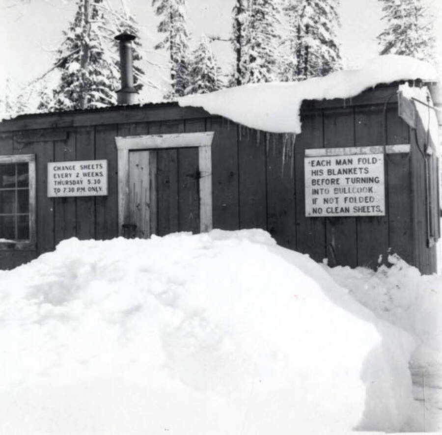 The laundry building at a logging camp in winter. The sign on the left reads ' Change sheets every 2 weeks . Thursday 5:30 to 7: 30pm only' The sign on the right reads 'Each man fold his blankets before turning into bllcook. If not folded no clean sheets.' Printed on the photograph is 'July 1965'