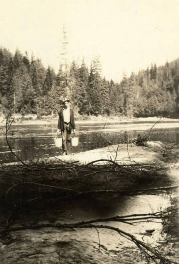 A man stands holding two buckets near a water source. Written on the back of the photograph is 'At the mouth of Beaver Cr. - on the North Fork of Clearwater - 1931.'
