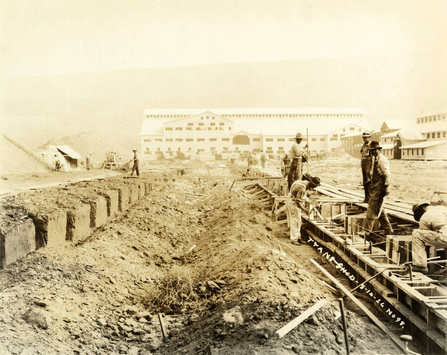 Men work on the foundation for one of the buildings at the Lewiston Mill. On the left side of the photograph are cement blocks. In the background is the mill. Written on the photograph is 'Trans-Shed 8/14/1926 No. 98'