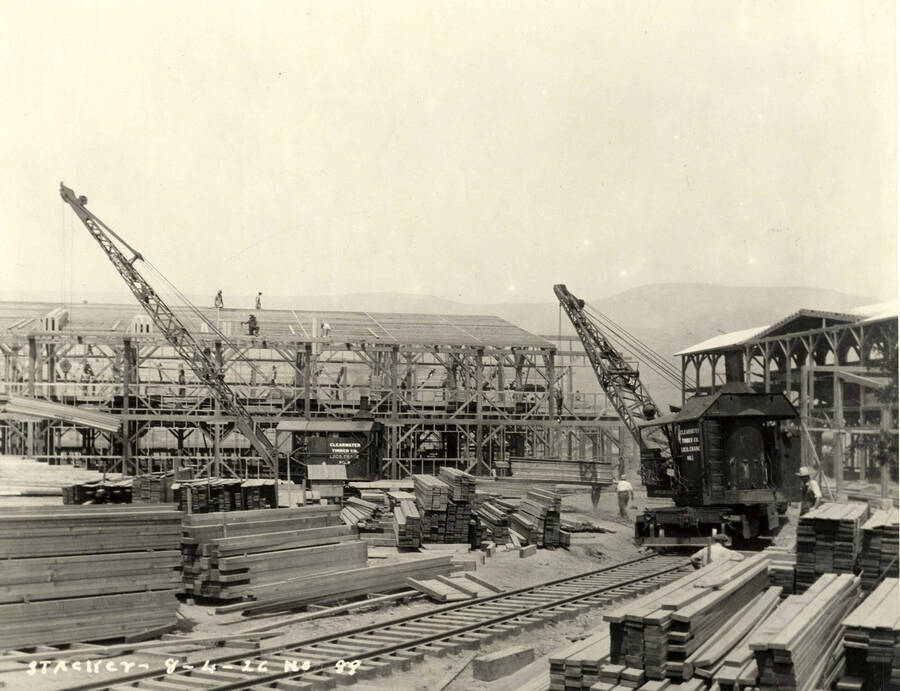 Two cranes, one a locomotive crane, work on buildings at the Lewiston Mill. Surrounding them are piles of lumber. Written on the photograph is 'Stacker 8/4/1926 No. 89'