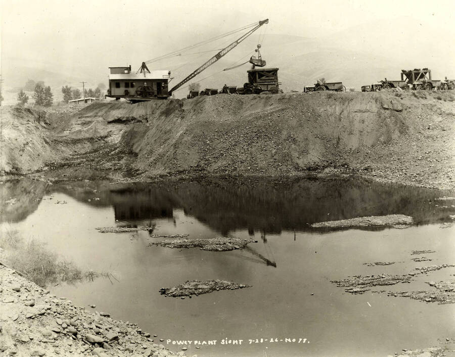 A crane dumps material into a dump area. In front of the crane is a pond-filled pit. Written on the photograph is 'Power Plant site 7/23/1926 No. 77'