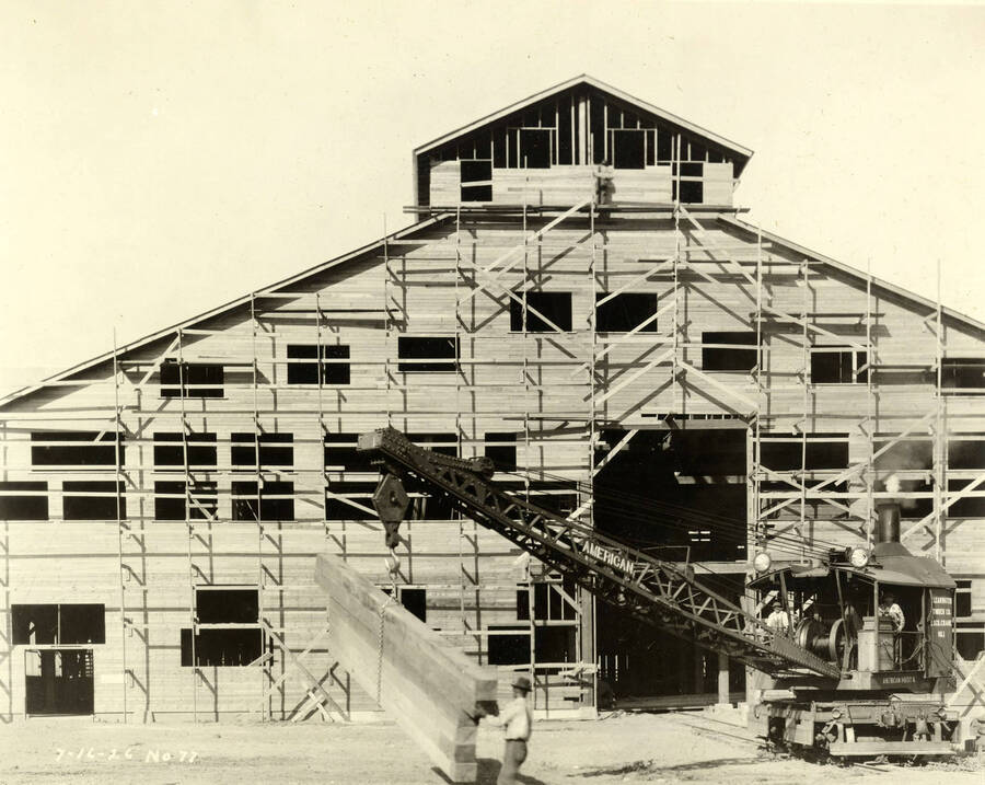 A crane lifts six pieces of lumber to help in building a building at the Lewiston Mill. A man helps move the set of lumber on the crane. A man stands at the top of the building on the scaffolding. Written on the photograph is '7/16/1926 No. 77'