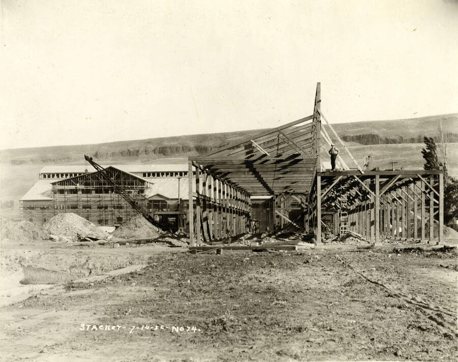 A man stands atop the framing for the stacker building while another building is being constructed in the background. Written on photograph is 'Stacker 7/14/1926 No. 74'