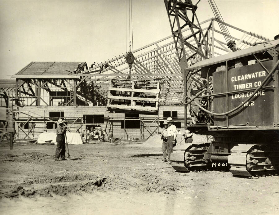 A crane lifts piece of machinery while three men stand underneath it. On the side of the crane, the writing reads 'Clearwater Timber Co. Loco. Crane No. 2.' Written on the photograph at the bottom of the crane is 'No. 61'.