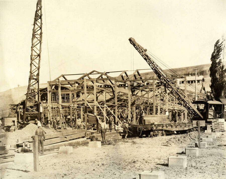 Men work to build the stacker building. The crane on the left hand side of the photograph is about pick up a load of lumber while the crane on the right hand side of the photograph is a locomotive crane and working with a man to attach lumber. Behind the cranes stands the framing for the building. Written on the photograph is 'Stacker Building 7/4/1926 No. 72'