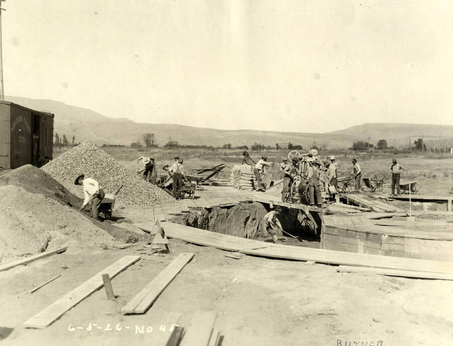 Men work to construct the burner at the Lewiston Mill. Piles of dirt and gravel sit at the left hand side of the photograph and men work on a piece of machine while men spread the material underneath them. Written on the photograph is 'Burner 6/5/1926 No. 45'
