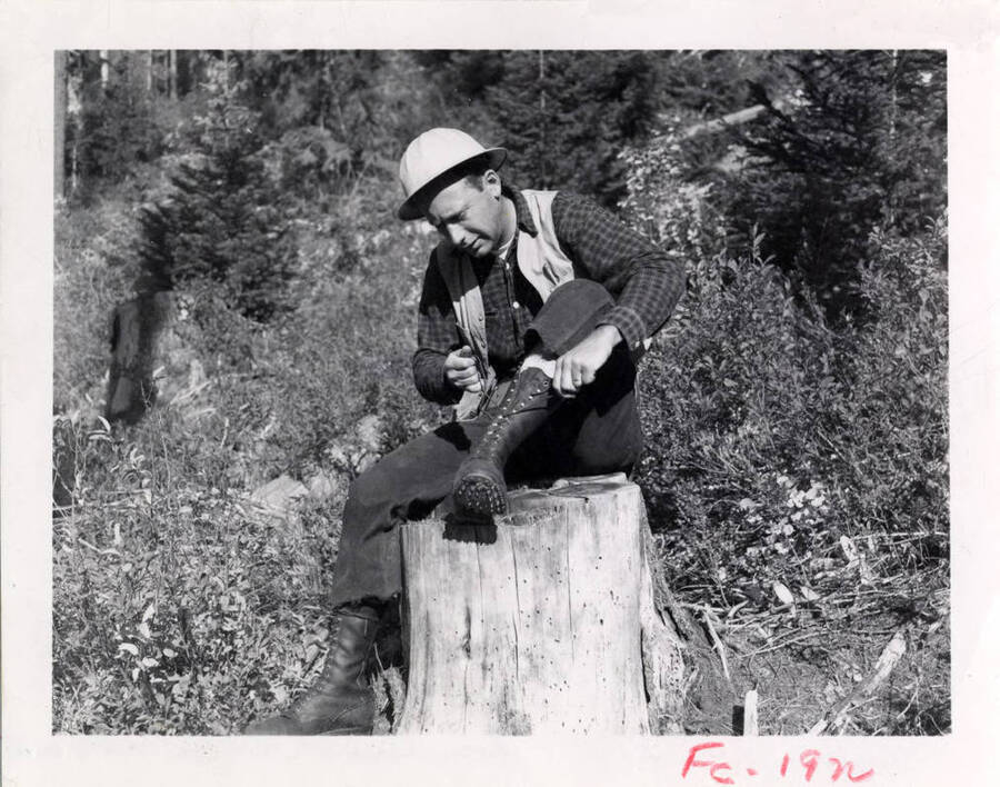 A forest worker laces up a boot while sitting on a stump. The description on the back says 'typical PFI foresters dress, note boots.' Stamped on the back is 'Photoby Jack M. Gruber Courtesy of Potlatch Forests, INC.'