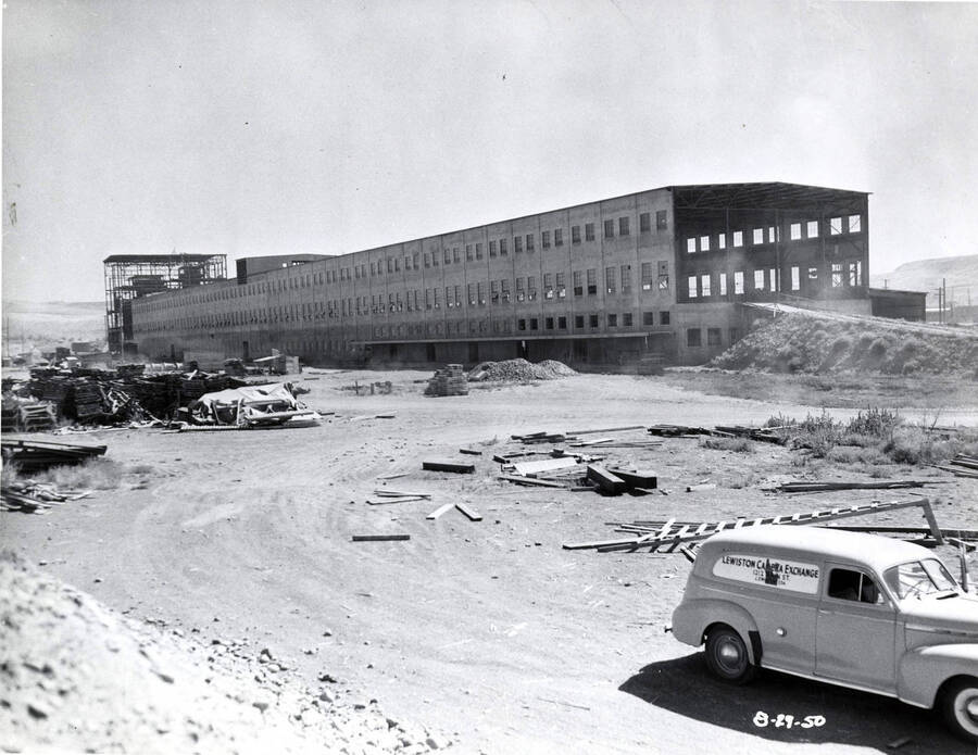 The outside frame of the Clearwater paper mill under construction. Also included in the picture is an automobile with a sign on it reading Lewiston Camera Exchange  1212 N. Main St, Lewiston, IDA.