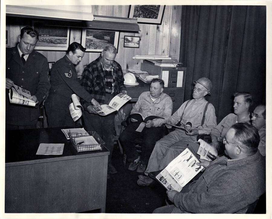 The description on the back of the photograph reads 'a typical training session on aircraft identification is held at Headquarters. Left to right at Sgt. George Webster, Sget. Gene Lookhart, James Delaney, Frank Steadman, Al Roeben, Headquarters area Skywatch supervisor for PFI; George Fraizer, Lawrence Baker and Bob Olin, mechanical superintendent of the woods.' Several of the men hold booklets that show different type of airplanes