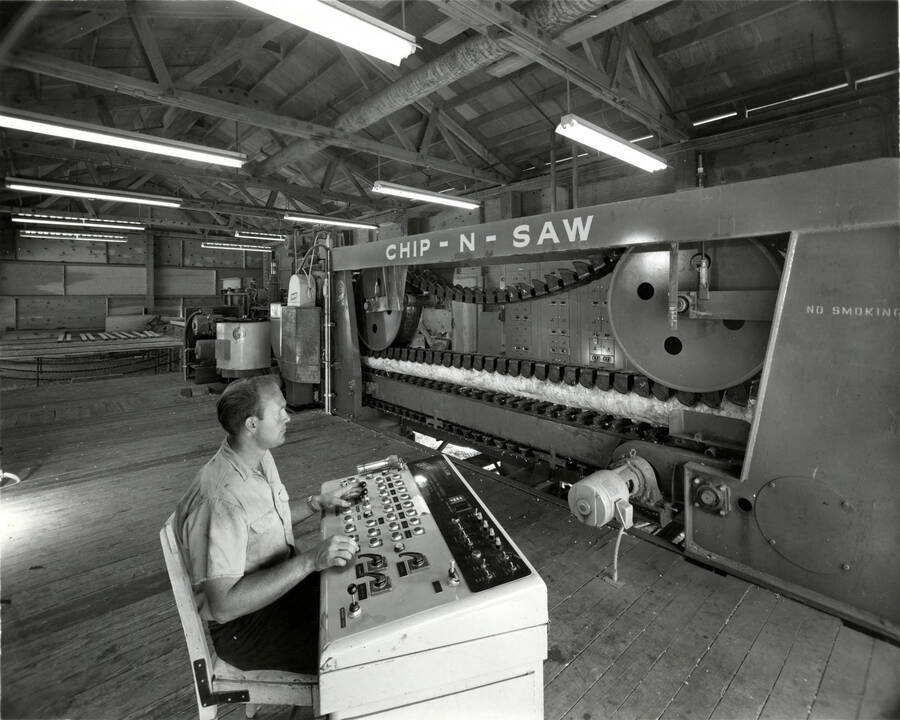 A man sits a control board. The description on the back reads: "Small log sawing equipment similar to that shown is in operation Potlatch Corporation's Lewiston Idaho sawmill. Double-cut band saws (head rigs) are also utilized there." There is a photography studio listed: Photographs by Richards Studio. Photographs since 1898. Pacific Ave. MA 7-9111. Tacoma, WAHS 98402.