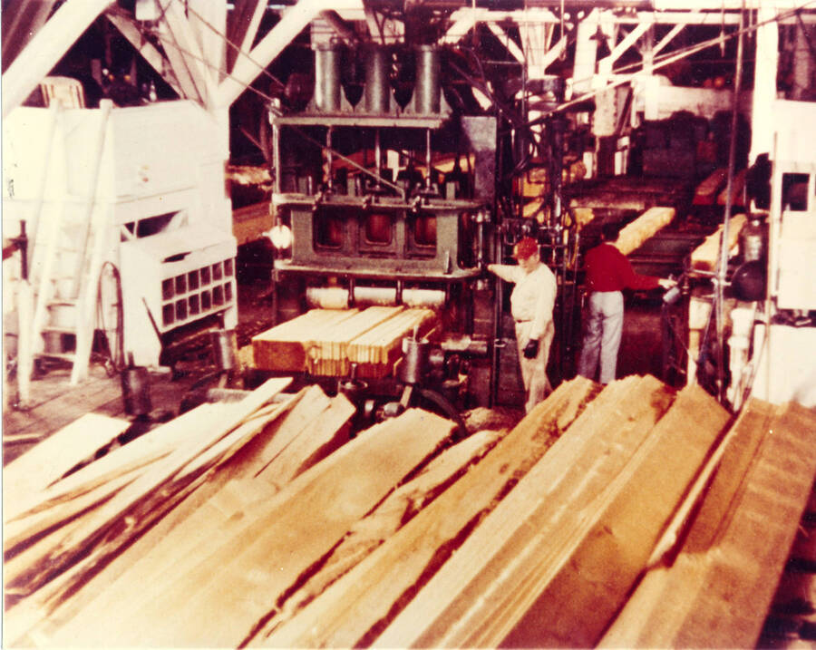 In the foreground, rough lumber waits to be processed. In the background, men work on planks.