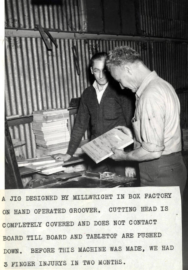 Two men work with boards of lumber. Attached to the photo is a description that reads "A jig designed by Millwright in box factory on hand operated groover. Cutting head is completely covered and does not contact board till board and tabletop are pushed down. before this machine was made we had 3 finger injurys in two months."