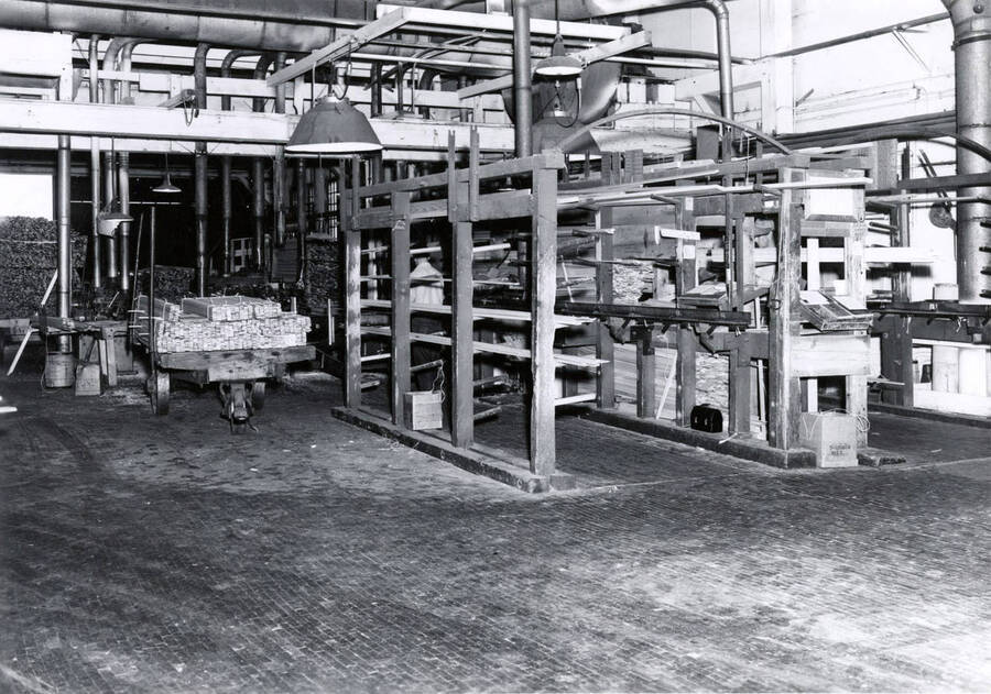 Bundles of wood boards sit waiting on a cart while others wait on racks to be put through the moulder, which is partially hidden by the racks. On the back, it is written "Moulders."