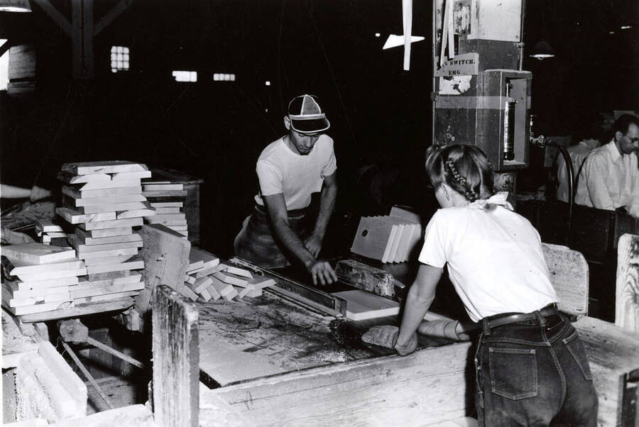A man and woman work together to run a piece of lumber through a machine. On the back, it's written "Box Fac."