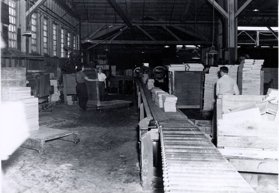 Men and women work together in the Clearwater Paper Mill. Stacks of planks of wood are on a roller conveyor belt as well as stacked on either side. On the back, it is labeled "workers in the box factory."
