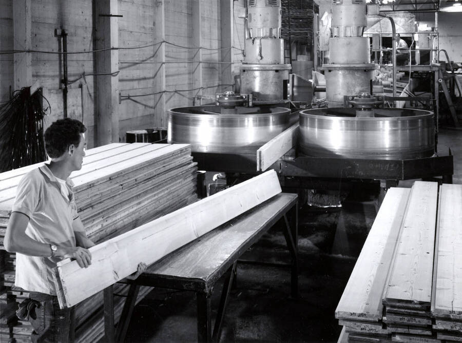 A man pushes a plank of wood through a machine. On the back, the description reads "Lock Deck laminated decking is manufactured at Potlatch Corporation's Lewiston, Idaho plant. This laminated product was developed by Potlatch and is used in the construction industry for both industrial and residential use. The product comes in a variety of sizes, grades and species." There is a stamp for Photographs by Richards Studios in Tacoma Washington on the back as well.