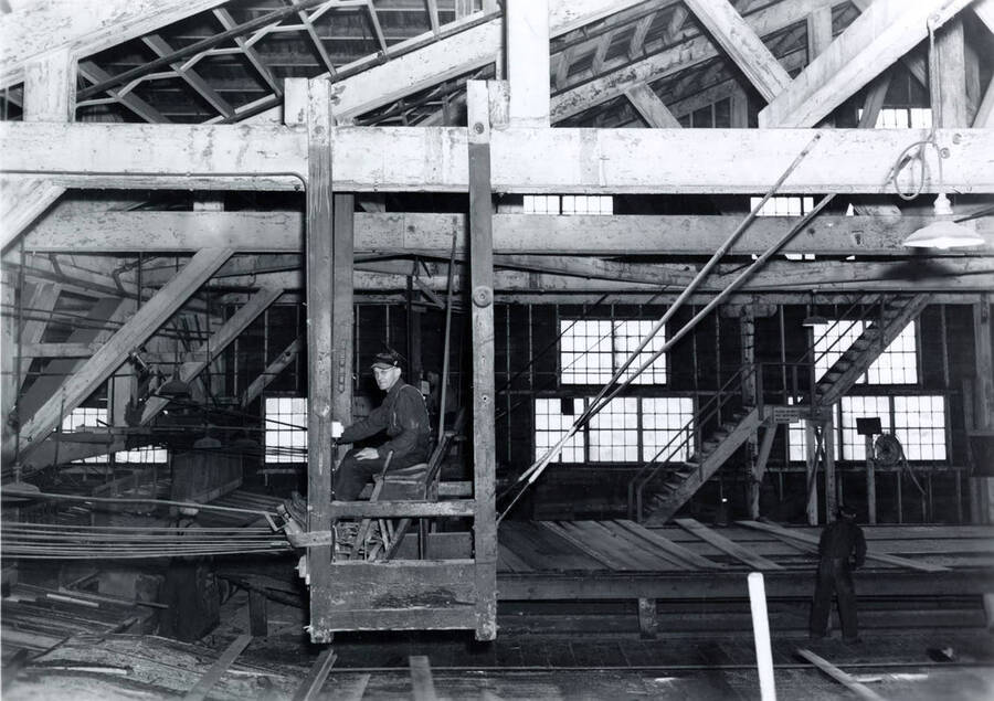 A man sits  in a chair  looking at the camera while men in the background work with planks of wood. The description on the back of photograph says "trimmer man."