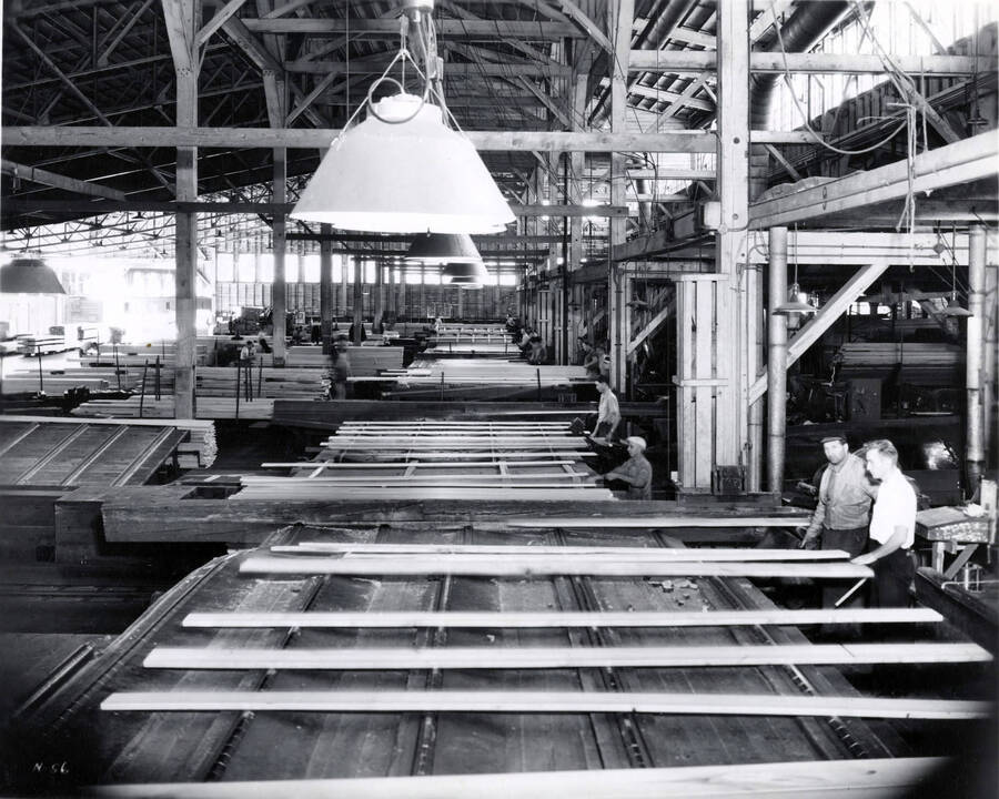 In the planer mill, planks of Lumber are inspected for grading. Two men stand on the right hand of the photo.