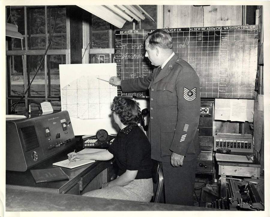The description on the back of the photograph reads "Sgt. George Webster, member of Air Force training team, explains use of map to Cora Todd, operator of radio at Headquarters. Blackboard in background is use to control movement of logging trains."